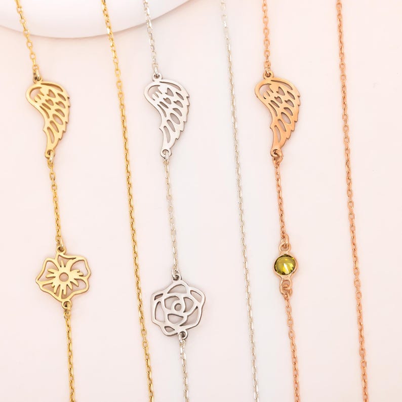 May include: Close-up of several delicate necklaces. One is gold with a wing charm and a flower pendant. Another is silver with a wing and rose charm. A third is rose gold with a wing and a green gemstone.