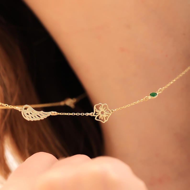 May include: A gold necklace with a wing charm, a flower pendant, and a small green gemstone. The necklace is worn around the neck, highlighting its delicate design. The chain is fine, and the overall aesthetic is minimalist. The necklace is made of gold.
