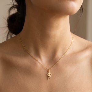 Op de afbeelding: Een delicate goudkleurige ketting met een kleine bladhanger. De hanger heeft een gedetailleerd, getextureerd ontwerp met meerdere kleine bladeren. De ketting wordt om de nek gedragen, met de ketting en hanger duidelijk zichtbaar.