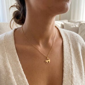 Op de afbeelding: Een goudkleurige ketting met een kattenhanger. De hanger is een silhouet van een kat met een kleine letter 'G' aan de zijkant. De ketting wordt gedragen door een persoon die een crèmekleurig vest draagt.