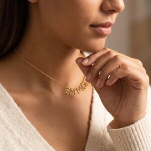 Può includere: Una collana color oro con il nome "EMMA" scritto in lettere maiuscole. La collana è indossata intorno al collo, con le lettere appese in una linea curva. La catena e le lettere sembrano essere di metallo lucido.