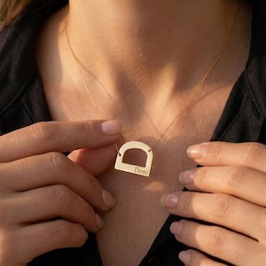 Op de afbeelding: Een goudkleurige ketting met een hanger in de vorm van een afgeronde boog. De hanger is gegraveerd met de naam "Demi". De ketting wordt om de nek gedragen.