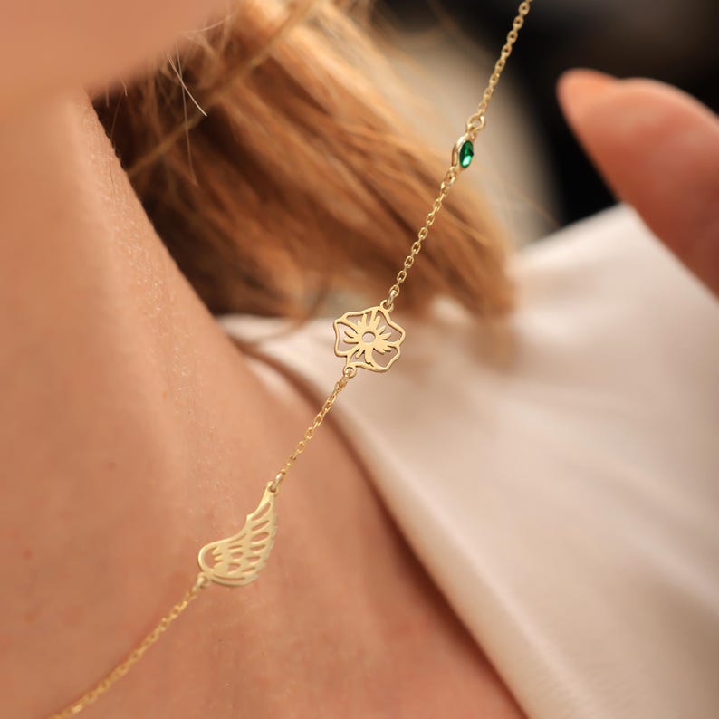 May include: A delicate gold necklace with a small wing charm, a stylized flower, and a green gemstone. The necklace is shown against a soft, neutral background, highlighting its intricate design and fine details. The chain is thin and elegant.