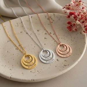 Può includere: Tre collane personalizzate con ciondoli circolari a strati. Le collane sono in oro, argento e oro rosa. Ogni ciondolo è inciso con i nomi Randy, Tiffany e Frances. Le collane sono esposte su una superficie color crema.