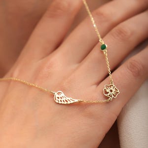 May include: A delicate gold necklace featuring a wing charm, a flower pendant, and a small green gemstone. The necklace is displayed on a hand, showcasing its intricate design and fine details. The jewelry is a stylish accessory.