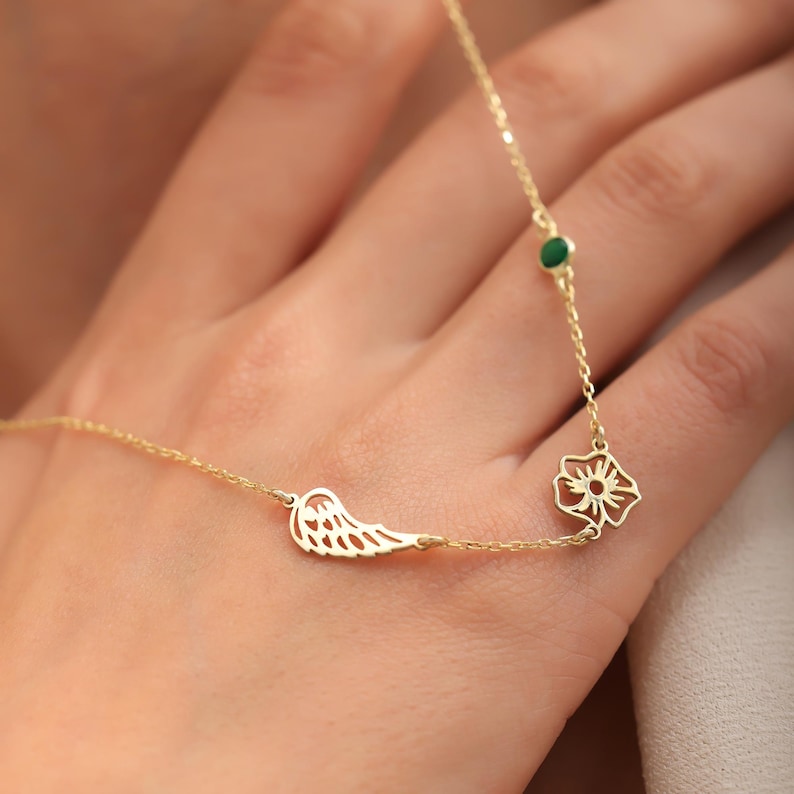 May include: A delicate gold necklace featuring a wing charm, a flower pendant, and a small green gemstone. The necklace is displayed on a hand, showcasing its intricate design and fine details. The jewelry is a stylish accessory.