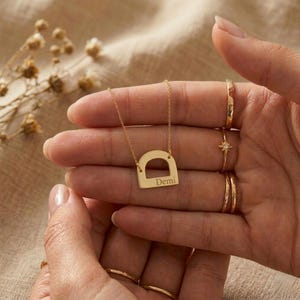 Op de afbeelding: Een goudkleurige ketting met een hanger in de vorm van een hoofdletter "D" met de naam "Demi" gegraveerd. De ketting wordt vastgehouden door een hand, en er zijn verschillende gouden ringen op de vingers te zien.