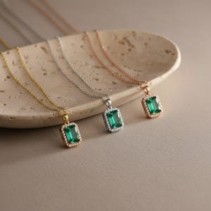 Op de afbeelding: Halskettingen met smaragdgroene edelsteenhangers met smaragdgroene slijpvorm. De hangers zijn omgeven door kleine, heldere stenen en gezet op gouden, zilveren en roségouden kettingen. De sieraden worden gepresenteerd op een beige, getextureerd oppervlak.