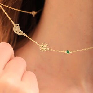 May include: A gold necklace with a wing charm, a flower pendant, and a small green gemstone. The necklace is worn around the neck. The chain is visible. The design is simple and elegant, suitable for everyday wear.