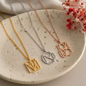 Op de afbeelding: Drie kettingen met dunne metalen schakels en geometrische hangers. Eén hanger is een goudkleurig vierkant met lineaire uitsparingen, één is een zilverkleurige cirkel met interne lijnen en één is een roségoudkleurige rechthoek met opengewerkt design.