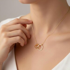 Op de afbeelding: Een goudkleurige ketting met twee in elkaar grijpende ringen. Eén ring is effen, de andere heeft gegraveerde tekst. De ketting hangt aan een fijne ketting. De sieraden worden getoond op de nek van een persoon.