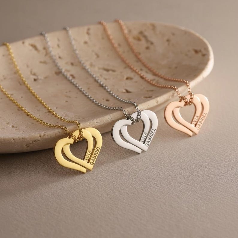 Op de afbeelding: Drie kettingen met hartvormige hangers in goud-, zilver- en ros&eacute;goudtinten. Elke hanger is voorzien van twee in elkaar grijpende hartcontouren met de namen IVIA en GABRIEL verticaal gegraveerd aan de zijkant van het hartframe.