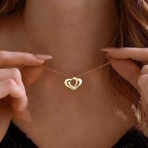 Könnte beinhalten: Eine goldfarbene Halskette mit zwei ineinander verschlungenen Herzanhängern. Die Anhänger sind durch eine feine Kette verbunden. Die Halskette wird von einer Person gehalten.