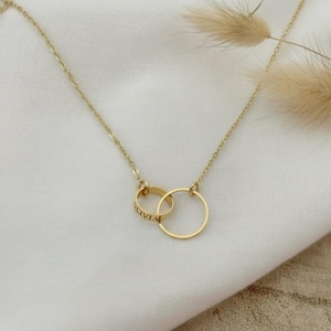 Op de afbeelding: Een goudkleurige ketting met twee in elkaar grijpende cirkels. Eén cirkel is gegraveerd met de naam "OLIVIA". De ketting wordt gepresenteerd op een wit oppervlak, met een zachte, neutrale achtergrond.