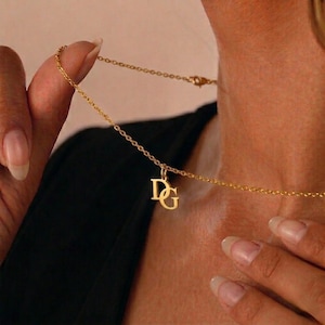 May include: A gold necklace with a fine chain and a pendant displaying the intertwined letters "DG." The necklace is shown against a dark backdrop, emphasizing its elegant design. The pendant is approximately 2 cm in height.
