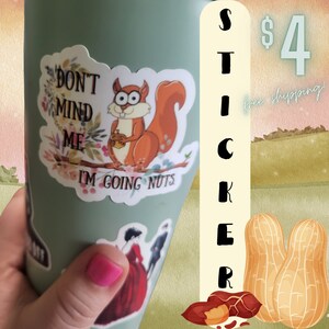 May include: A green travel mug with a sticker of a cartoon squirrel holding a nut and the text "Don't mind me I'm going nuts". The sticker is on a green travel mug. The mug is being held by a hand with pink nail polish. The background is a blurred image of a field with a sign that says "Sticker $4 free shipping!"