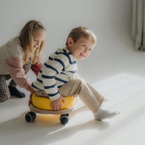 Puede incluir: Un niño pequeño está sentado en un juguete con ruedas amarillo y de madera, con un suéter a rayas azules y blancas y pantalones color caqui. Una niña con una blusa rosa y beige y pantalones con estampado de leopardo lo ayuda. El juguete tiene cuatro ruedas negras.