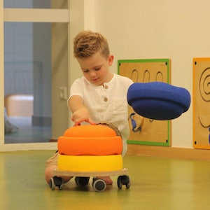 Peut inclure: Un enfant joue avec un chariot jouet coloré. Le chariot a trois pièces rondes empilées en mousse, jaunes, oranges et bleues. Le chariot a quatre petites roues. L'enfant porte une chemise blanche.