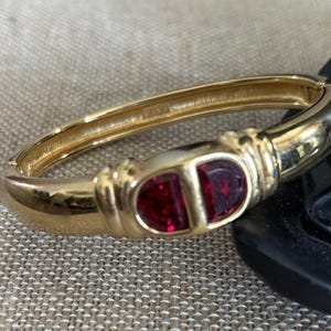May include: A gold-toned hinged bangle bracelet with a rectangular red jewel at the center. The bracelet has a smooth, polished finish and a classic design. The red jewel is divided into two sections.