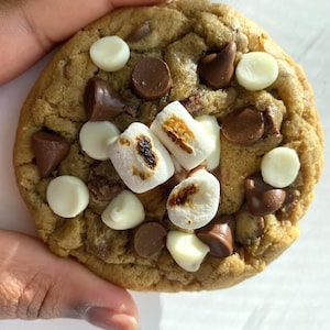 May include: A large, round cookie with a golden-brown exterior. The cookie is studded with white chocolate chips, milk chocolate chips, and toasted marshmallows. The cookie is held by a hand.