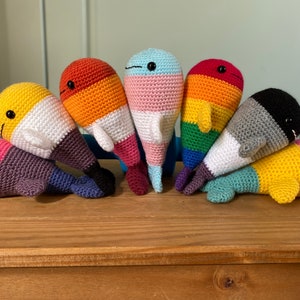 May include: Seven crocheted whale plushies in a rainbow of colours, including pink, yellow, orange, white, blue, green, and black. Each whale has a different colour pattern and is arranged in a semi-circle.