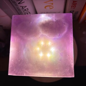 May include: A square, translucent object with a galaxy-like design. The object has a pinkish-purple hue with a bright yellow centre. The edges are sharp, and the overall effect is ethereal and artistic.