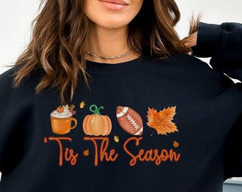 May include: Black sweatshirt with a graphic design of a pumpkin, a football, a cup of coffee, and leaves. The text "Tis the Season" is written in orange below the graphic.
