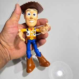 May include: A plastic toy figure of Woody from the Toy Story franchise. Woody is wearing his signature brown cowboy hat, yellow shirt, blue jeans, and brown boots. He is smiling and has his arms outstretched.