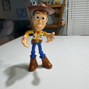 May include: A plastic toy figure of Woody from the Toy Story franchise. Woody is wearing his signature brown cowboy hat, yellow vest, blue jeans, and brown boots. He has a friendly expression on his face.