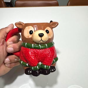 May include: A ceramic mug shaped like a bear, featuring a brown head with black eyes and a black nose. The bear wears a red sweater with green trim. The handle is red.
