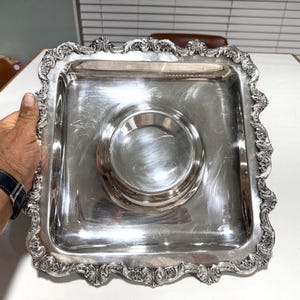 May include: A square, silver-colored serving tray with an ornate, scrolled edge. The tray has a circular indentation in the center. The tray is held by a person's hand, with a black watch visible on the wrist.