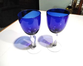 May include: Two cobalt blue wine glasses with clear stems and bases. The glasses are empty and sitting on a white surface.