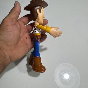 May include: A plastic toy figure of Woody from the Toy Story franchise. Woody is wearing his signature brown cowboy hat, blue jeans, and a yellow shirt with a red bandana and a sheriff's star. He is holding his right arm out, as if he is about to wave.