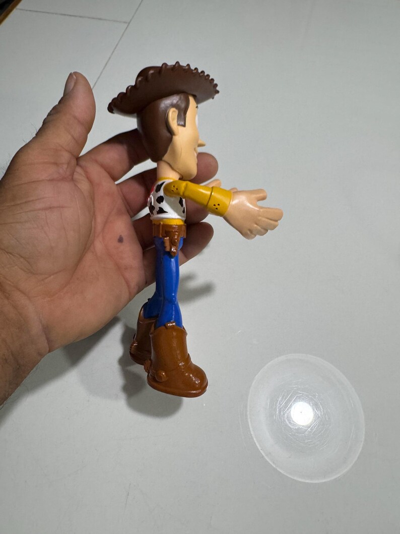 May include: A plastic toy figure of Woody from the Toy Story franchise. Woody is wearing his signature brown cowboy hat, blue jeans, and a yellow shirt with a red bandana and a sheriff's star. He is holding his right arm out, as if he is about to wave.