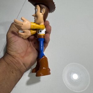 May include: A plastic toy figure of Woody from the Toy Story franchise. Woody is wearing his signature brown cowboy hat, yellow shirt, blue jeans, and brown boots. He is holding his arms out in front of him.