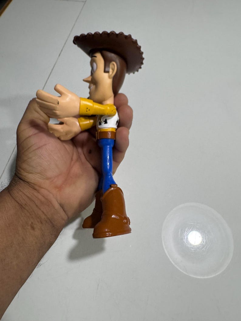 May include: A plastic toy figure of Woody from the Toy Story franchise. Woody is wearing his signature brown cowboy hat, yellow shirt, blue jeans, and brown boots. He is holding his arms out in front of him.