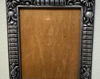 May include: A rectangular, ornate silver-colored picture frame. The frame has a detailed floral and scroll design around the edges and top. The center is a light brown wood. The frame is designed to hold a photograph or artwork.
