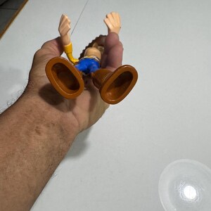 May include: A plastic toy figure of Woody from the Toy Story franchise. Woody is wearing his signature brown cowboy hat, yellow shirt, blue jeans, and brown boots. He is standing with his arms raised in the air.