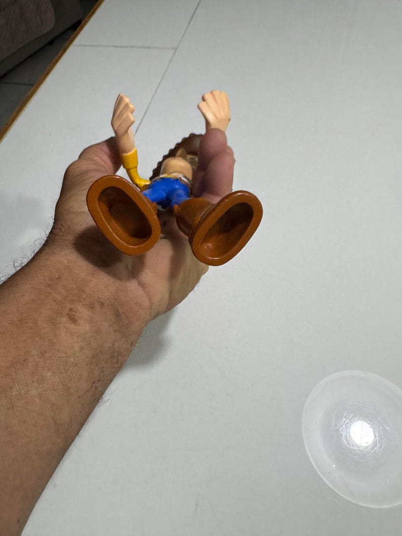 May include: A plastic toy figure of Woody from the Toy Story franchise. Woody is wearing his signature brown cowboy hat, yellow shirt, blue jeans, and brown boots. He is standing with his arms raised in the air.