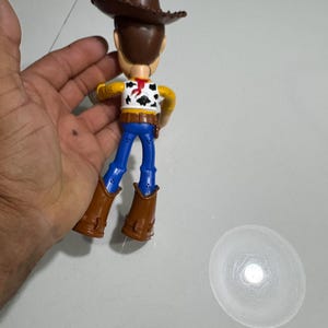 May include: A plastic toy figure of Woody from the Toy Story franchise. The figure is wearing a brown cowboy hat, a yellow shirt with a red bandana, blue jeans, and brown boots. The figure is standing on a white surface.