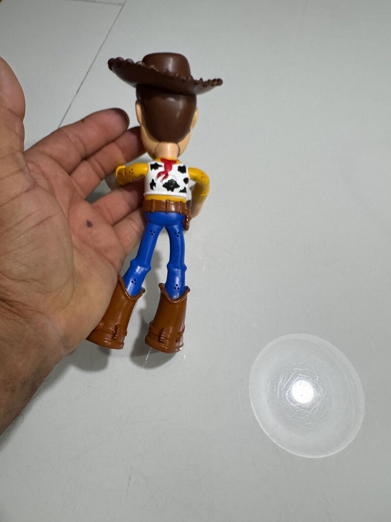 May include: A plastic toy figure of Woody from the Toy Story franchise. The figure is wearing a brown cowboy hat, a yellow shirt with a red bandana, blue jeans, and brown boots. The figure is standing on a white surface.