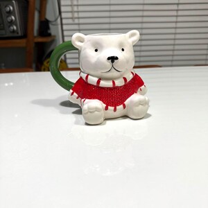May include: A ceramic polar bear mug with a red sweater and a green handle. The mug is white with black eyes and a black nose. The sweater has white stripes. The mug is designed to look like a bear sitting upright.