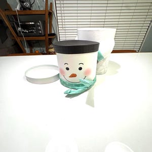 May include: A snowman-themed container with a black lid, a smiling face, and a teal scarf. The container is white with pink cheeks and black eyes. A white lid sits next to the container. A second white container is behind the snowman container.