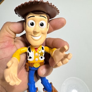 May include: A plastic toy figure of Woody from the Toy Story franchise. Woody is wearing his signature brown cowboy hat, yellow shirt with a sheriff's star, blue jeans, and brown boots. He has a friendly smile and is holding out his right hand.