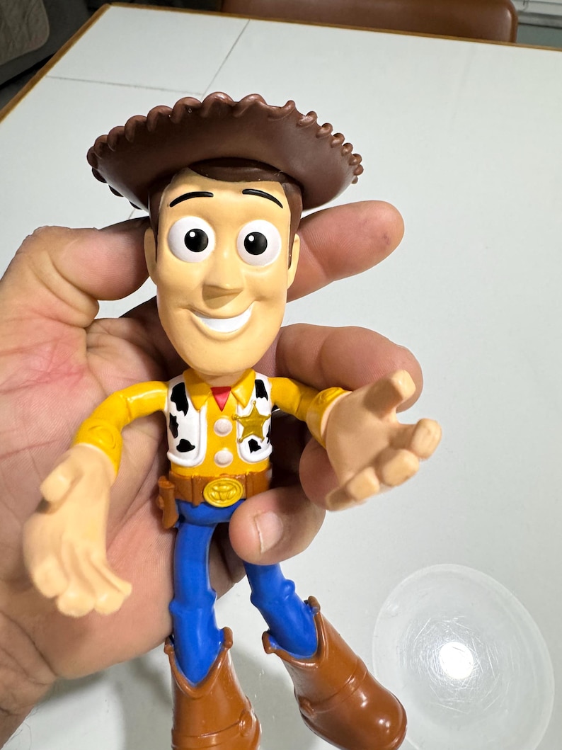 May include: A plastic toy figure of Woody from the Toy Story franchise. Woody is wearing his signature brown cowboy hat, yellow shirt with a sheriff's star, blue jeans, and brown boots. He has a friendly smile and is holding out his right hand.