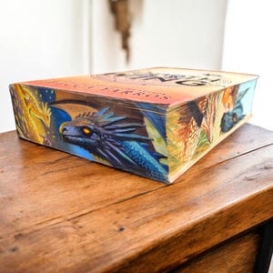 Op de afbeelding: Een hardcover boek met een drakenillustratie op de omslag. De draak is blauw met gele ogen en staat tegen een kleurrijke achtergrond. Het boek ligt op een houten oppervlak.