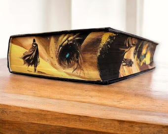 May include: A decorative book with a unique illustration on the edges. The artwork features a figure in a cape walking towards a large eye with a blue iris. The book has a black cover and is resting on a wooden surface.