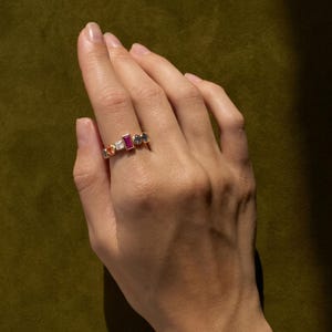May include: A gold ring with a series of colorful gemstones, including red, orange, white, and blue. The ring is set against a green background. The gemstones are rectangular and round, creating a unique design.