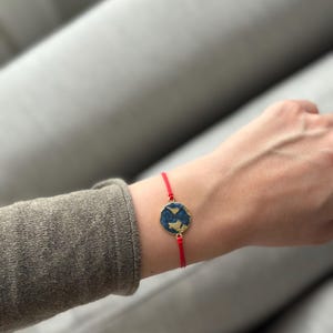 Könnte beinhalten: Ein rotes Armband mit einem goldfarbenen Anhänger, der eine blaue und goldene Weltkarte zeigt.