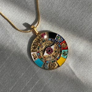 Pode incluir: Colar de ouro com um pingente redondo que apresenta um design de olho colorido cercado por vários símbolos e pedras preciosas.